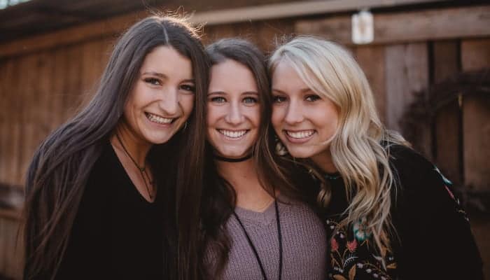 Two brunette women and one blonde woman stand with their heads together smiling in front of a wooden building