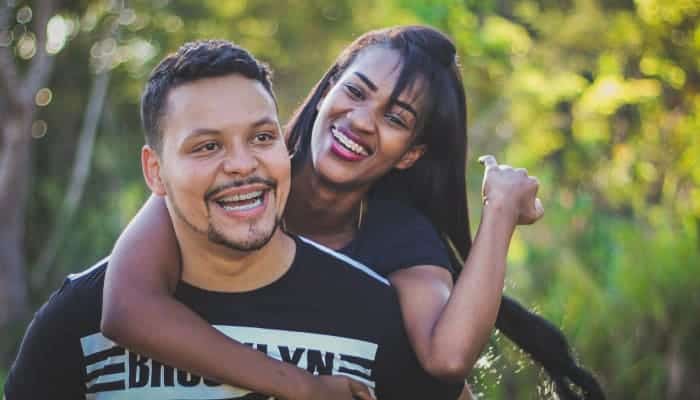 Dark-haired woman embracing a man from the back as they both smile while wearing braces outside in front of trees