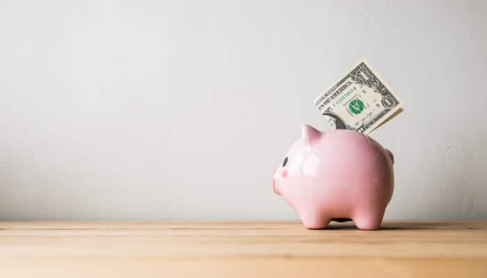 On a tan countertop, a green one dollar bill sits on top of a pink piggy bank used for saving money