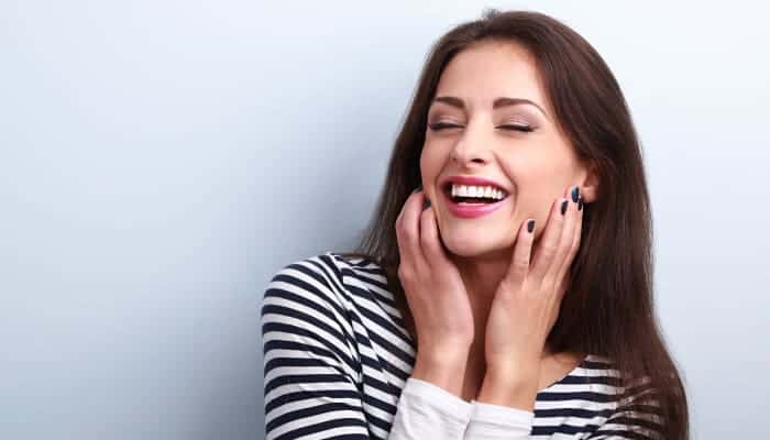 Brunette woman wearing permanent dental veneers and a striped shirt smiles as she touches her cheeks