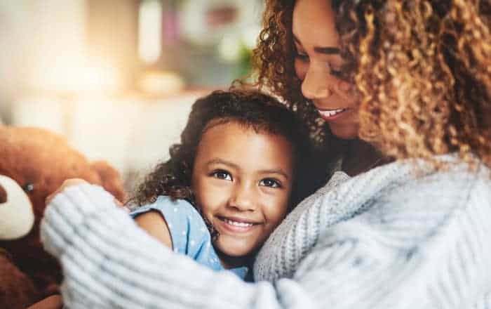 Curly-haired mother in a light gray sweater embraces her daughter who still has baby teeth while sitting on a couch